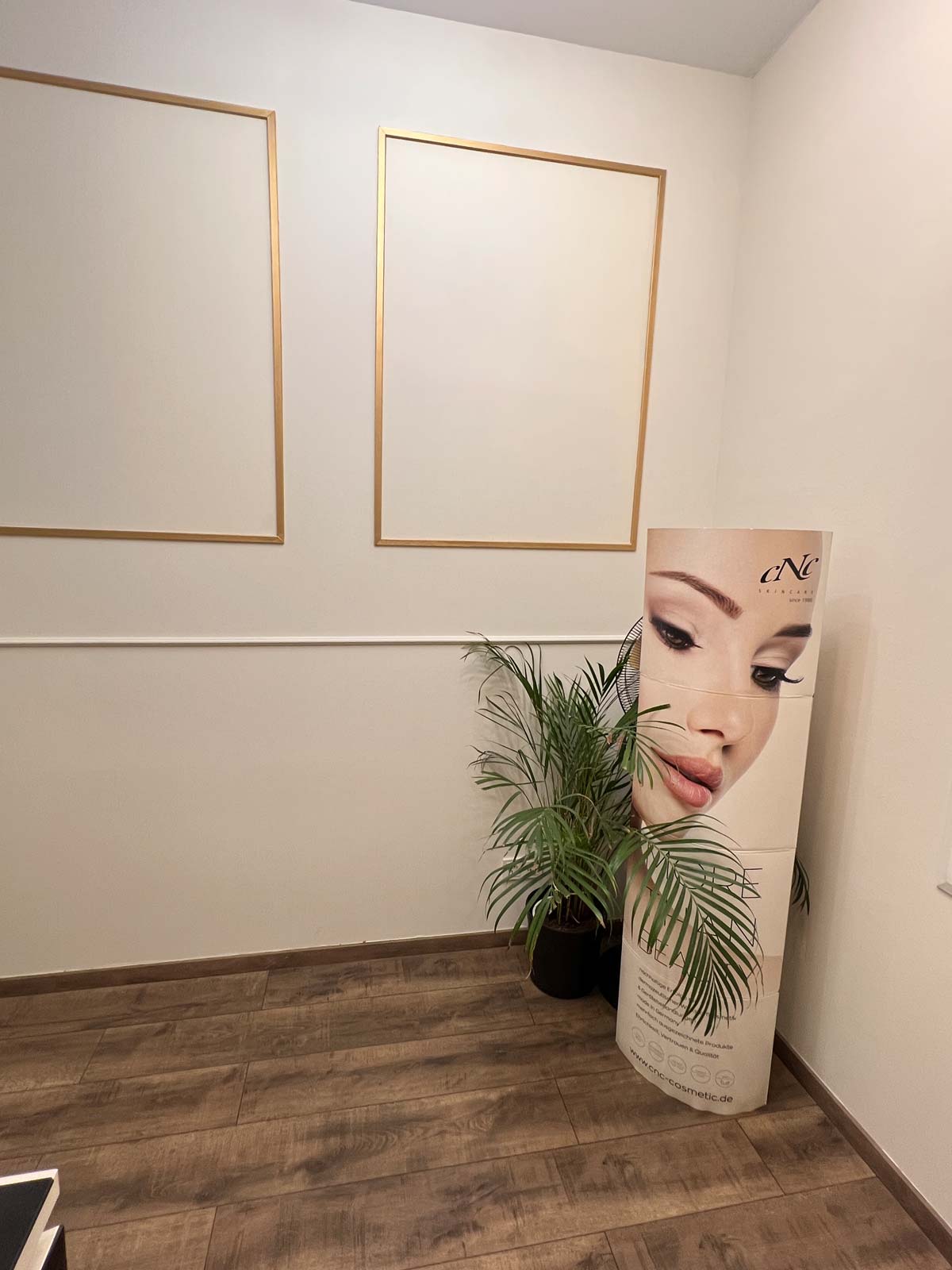 Leeres Zimmer mit Holzfußboden, zwei goldene Rahmen, Pflanze, und einem Werbeplakat mit Frauengesicht.
