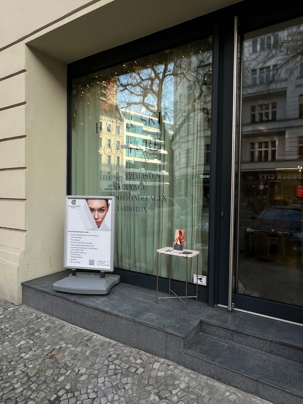 Geschäftseingang mit Werbeaufsteller, Glastür, Spiegelung von Bäumen und Gebäuden im Fenster.